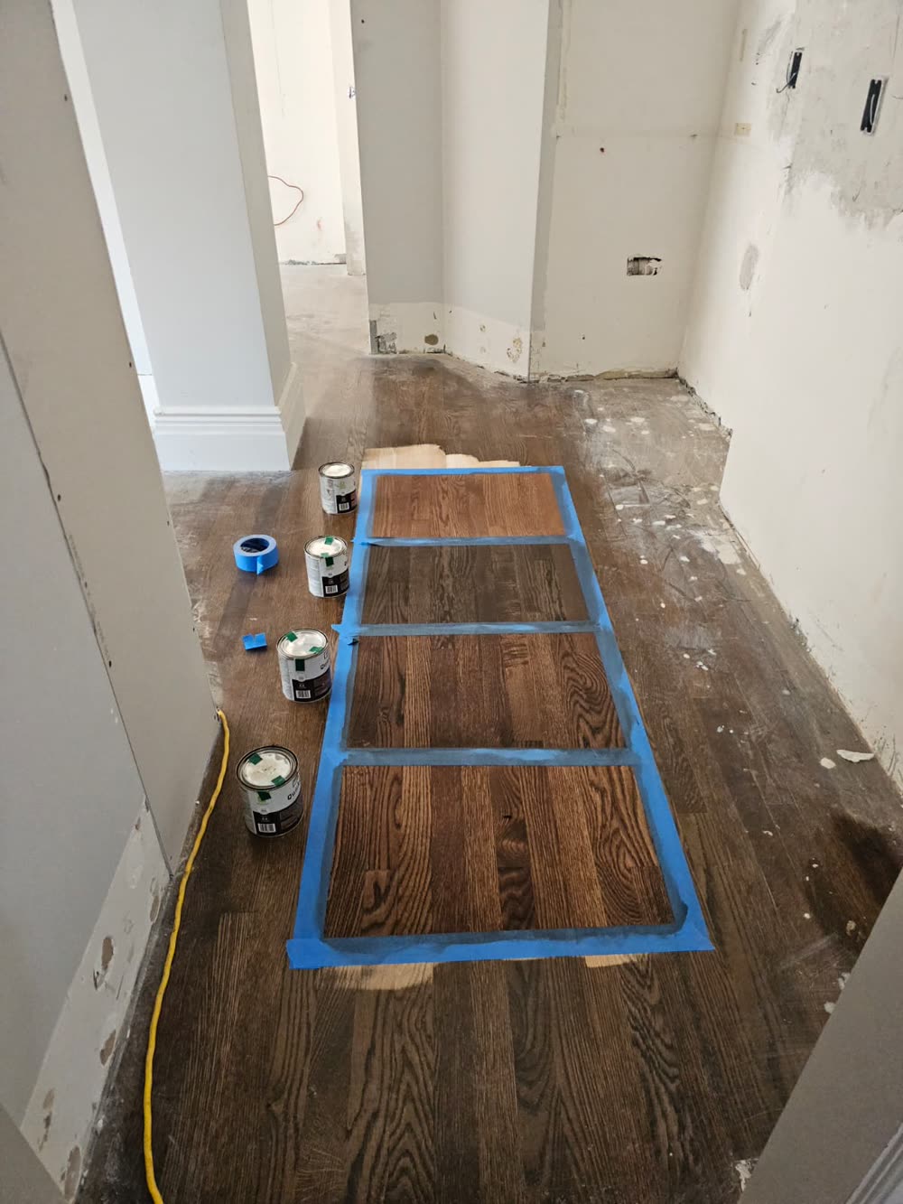Four stain samples applied side by side on an oak floor and taped off with blue painter's tape, with the stain cans next to them